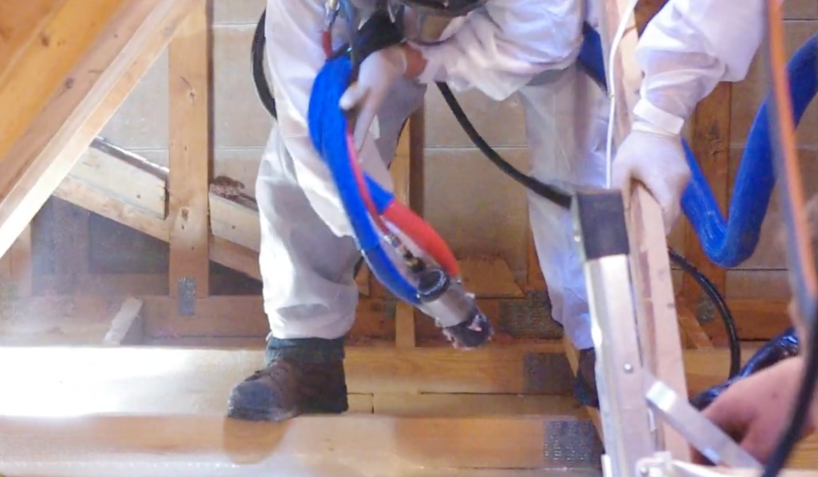 A worker in protective white coveralls sprays foam insulation into an attic space using a professional applicator gun.