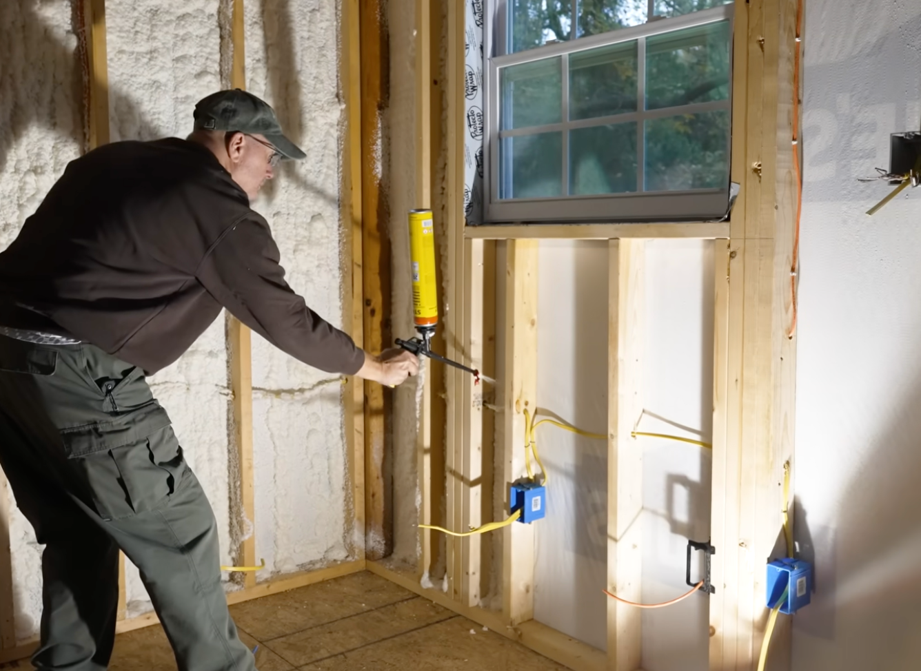 A person uses a spray foam gun to seal gaps in the wooden wall frame of a room under construction with electrical boxes.
