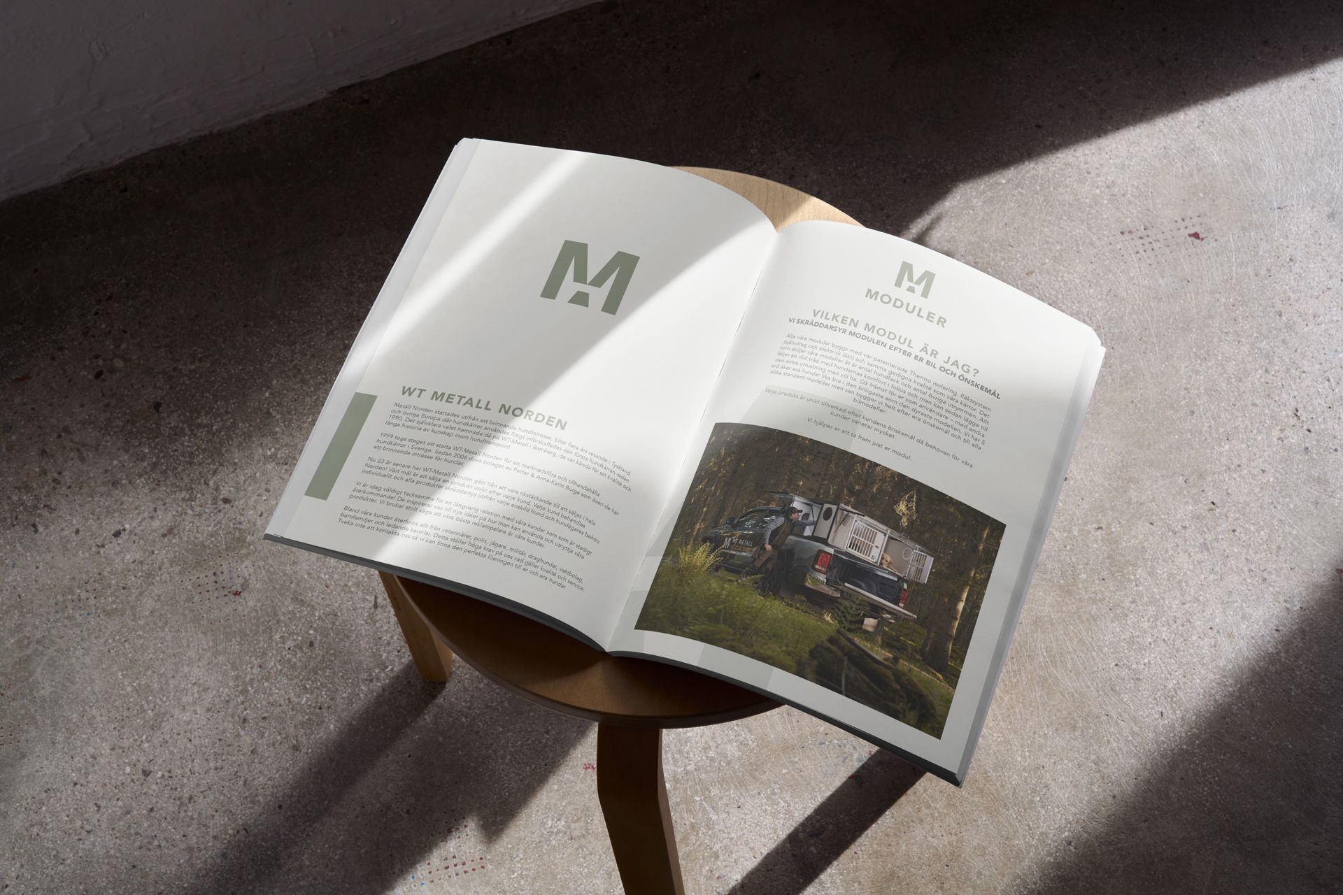 Bild på företagets produktkatalog fotad rakt ovanifrån. Katalogen ligger uppslagen på en rund träpall som står på ett betonggolv. Från höger övre hörn sipprar solljus in som skapar skuggor från pallen.
