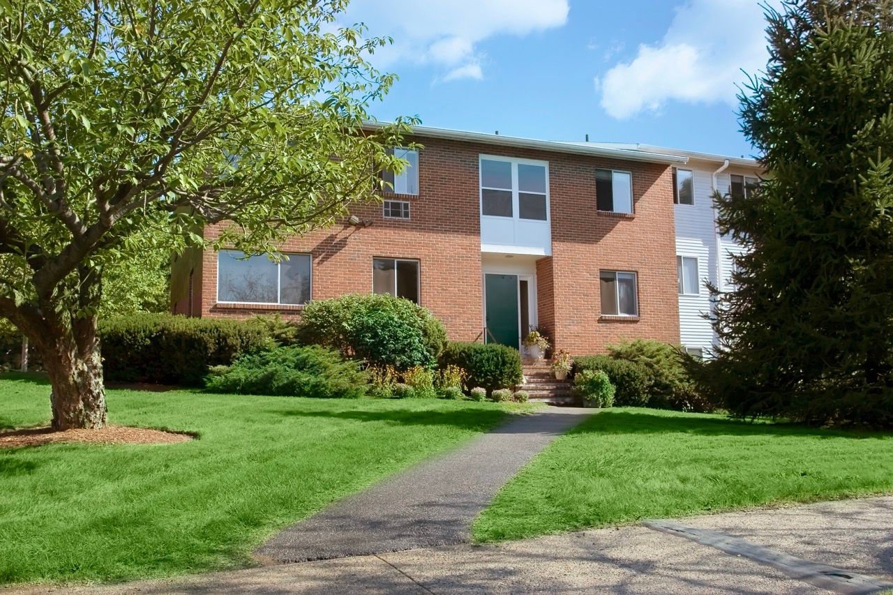 Exterior view of a brick apartment building with a landscaped lawn and walkway.
