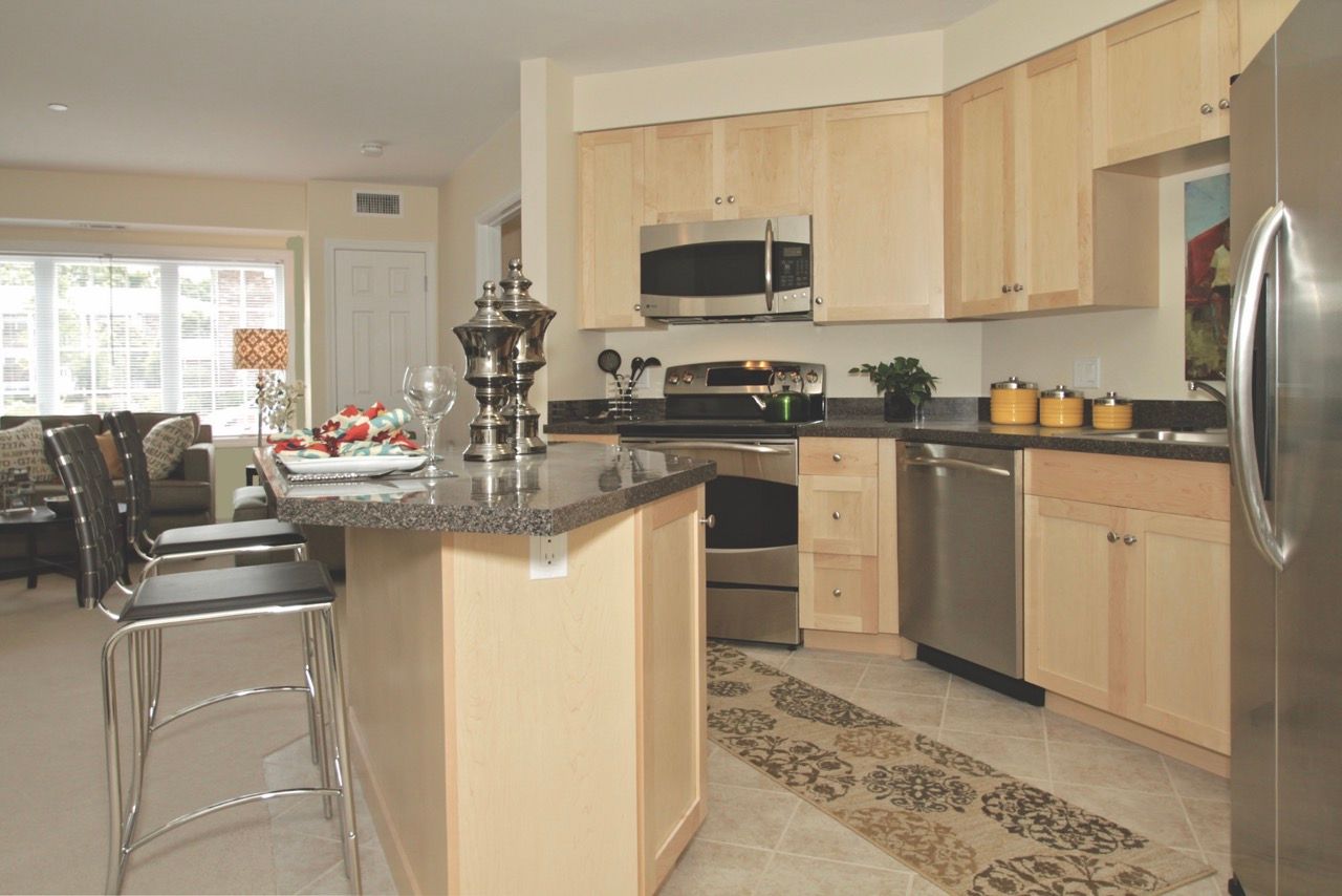 Open-concept apartment kitchen with light wood cabinets, stainless appliances, and a granite island.