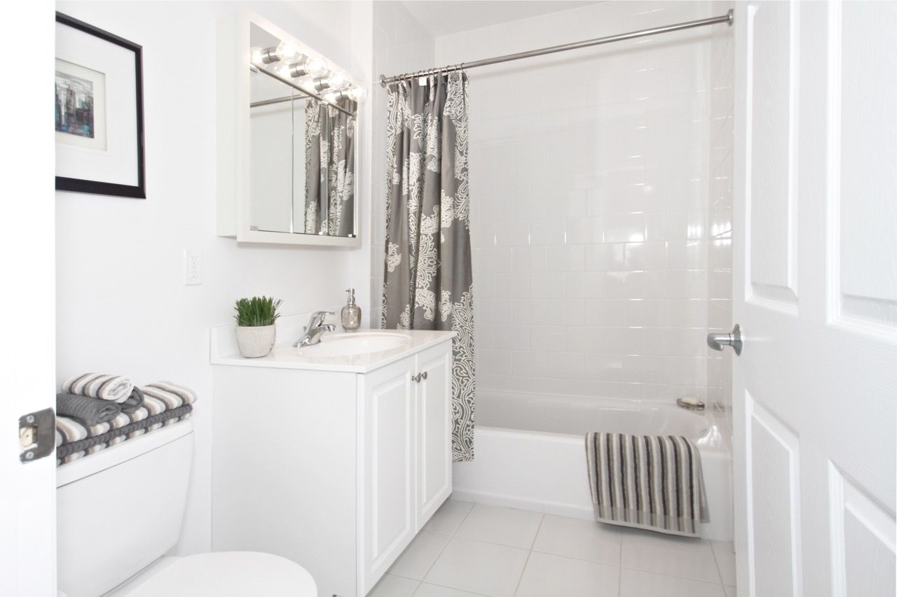 White apartment bathroom with vanity, mirror, toilet, and a tiled tub with a patterned shower curtain.
