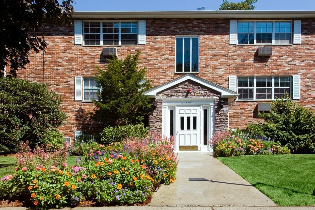 Exterior view of a brick apartment building entrance with a stone facade and colorful flower beds.