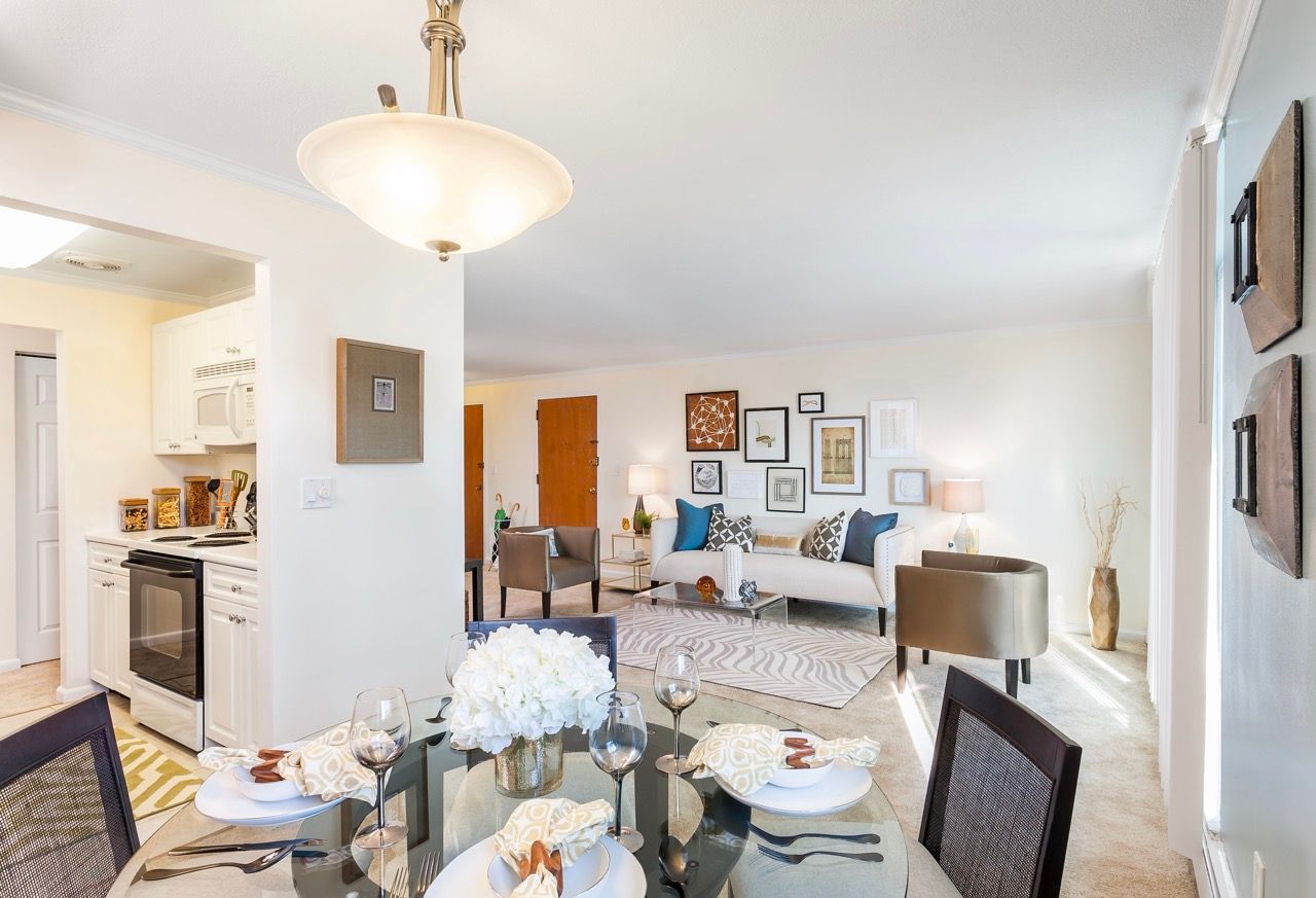 Open-concept living/dining area in an apartment with white kitchen, glass dining table, and gallery wall.