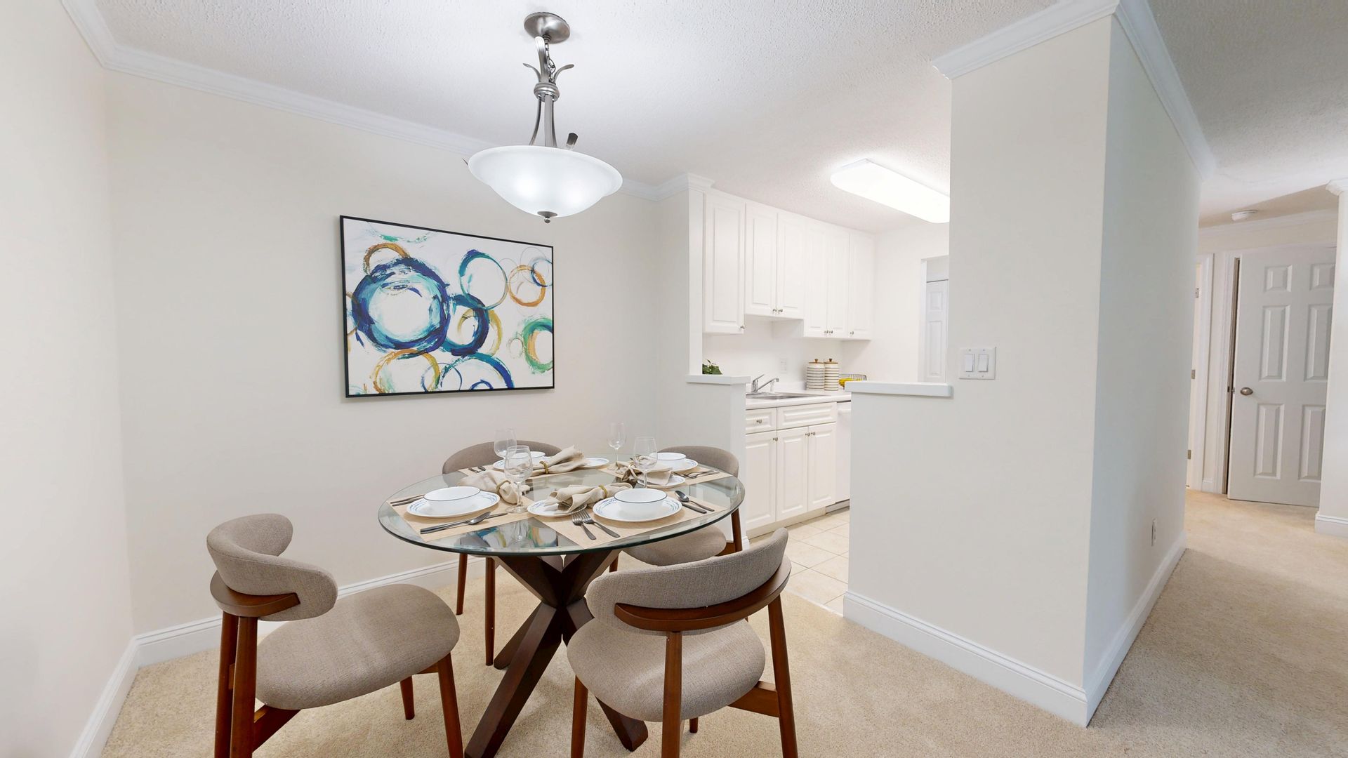 Dining area with round glass table, two chairs, abstract wall art, and a view into a bright kitchen and hallway.