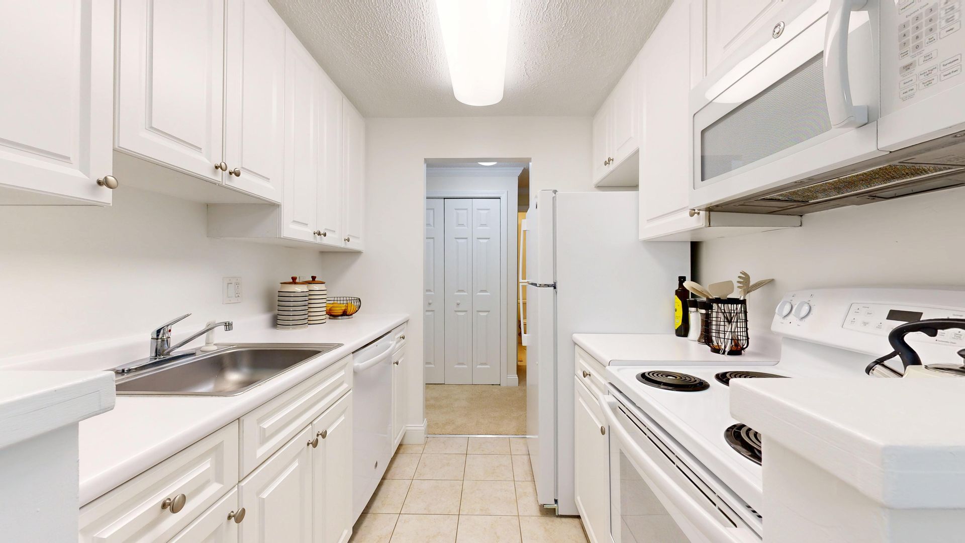 Bright white galley kitchen with cabinets, stove, sink, and a doorway at the far end.