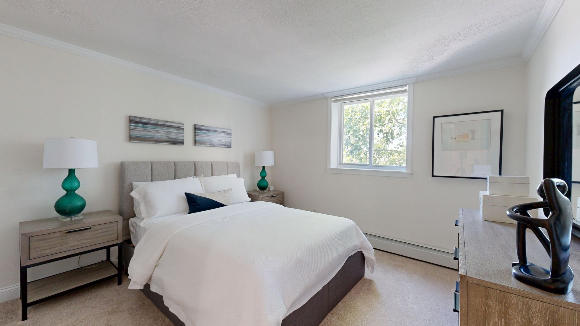 Bright bedroom with a neatly made bed, teal lamps, wall art, and a window.