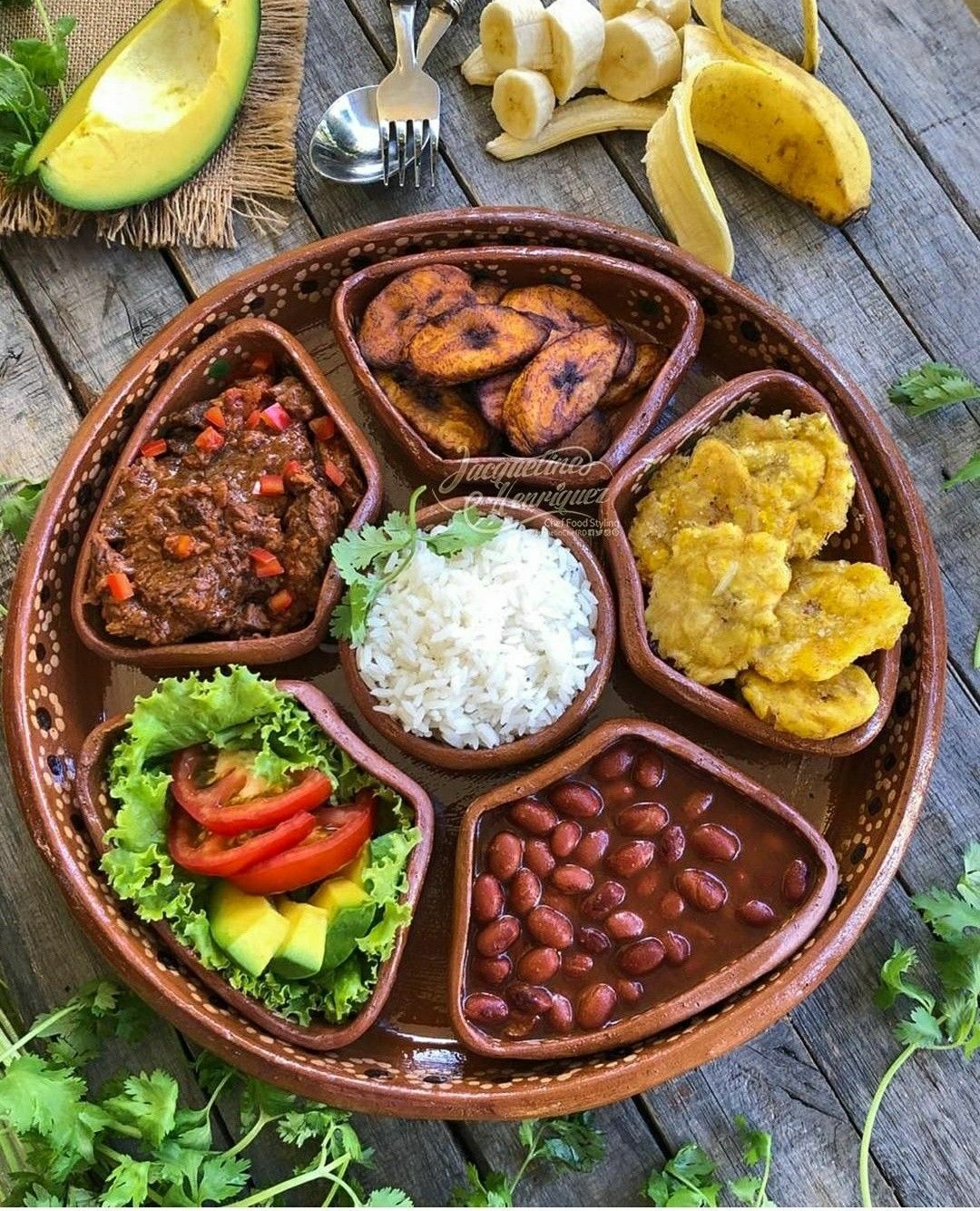 Round platter with various Latin American dishes: beef, fried plantains, beans, rice, salad, and tostones.