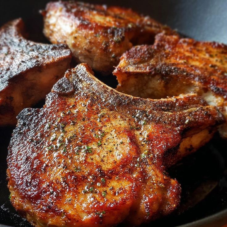 Close-up of several seared pork chops in a pan; golden-brown crust with visible herb seasoning.