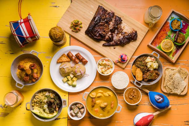Overhead shot of various Caribbean dishes on a colorful wooden table. Includes meat, stews, sides, and drinks.