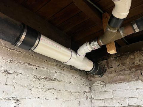 Exposed white PVC pipes and copper plumbing run along rough brick walls in a basement ceiling.