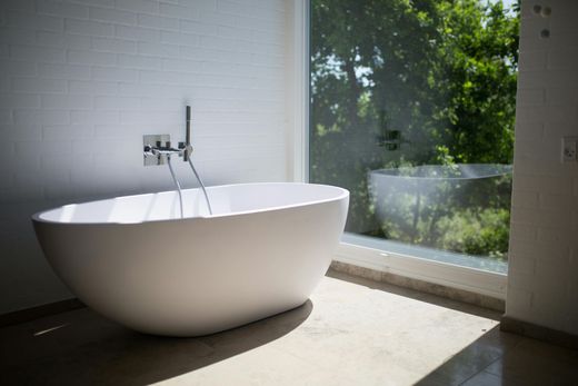 Modern bathroom with a white freestanding bathtub beside a large sunlit window overlooking greenery