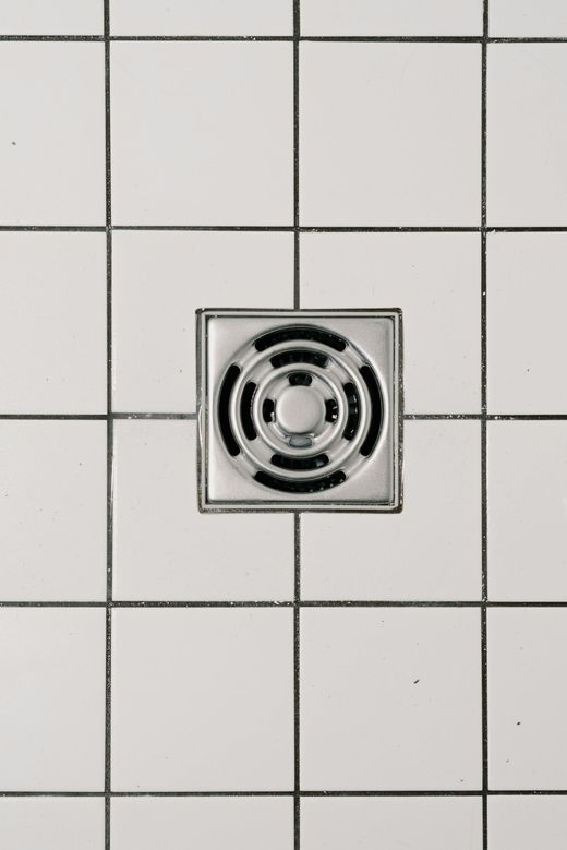 Square metal floor drain centered in white tiled flooring.