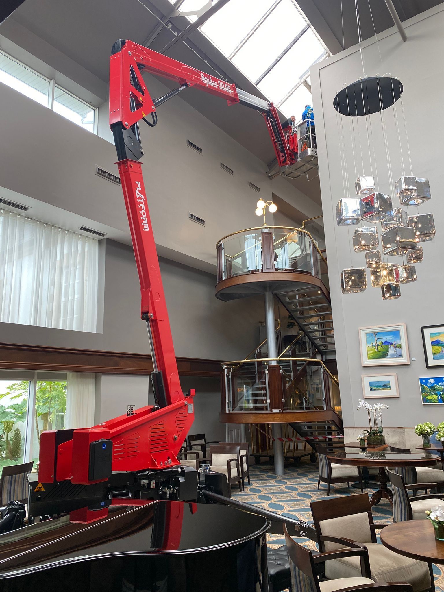 Red hydraulic boom lift in a bright modern atrium with high ceilings and hanging sculptures.