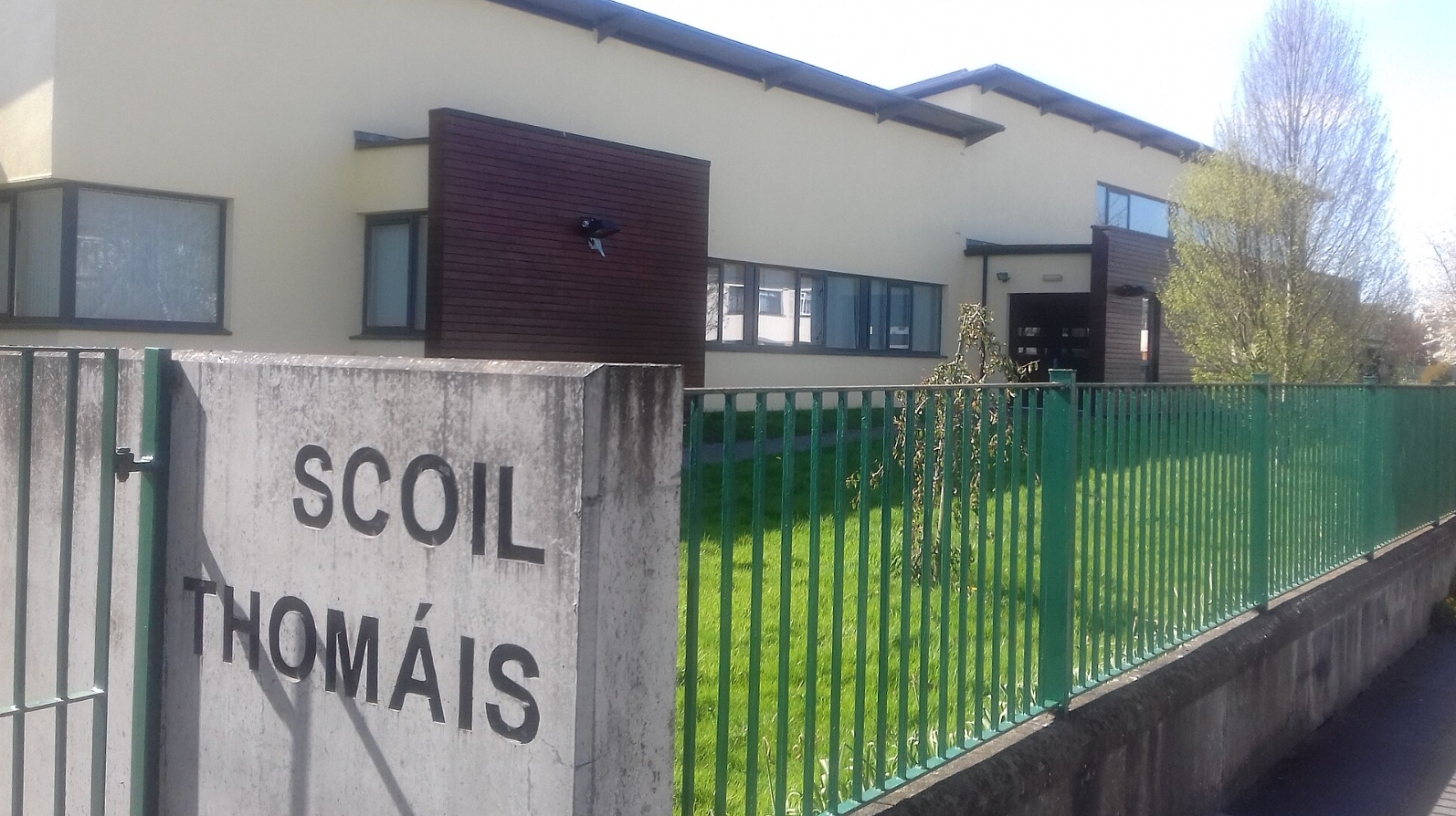 School building behind a green fence with a stone sign reading “SCOIL THOMÁIS”