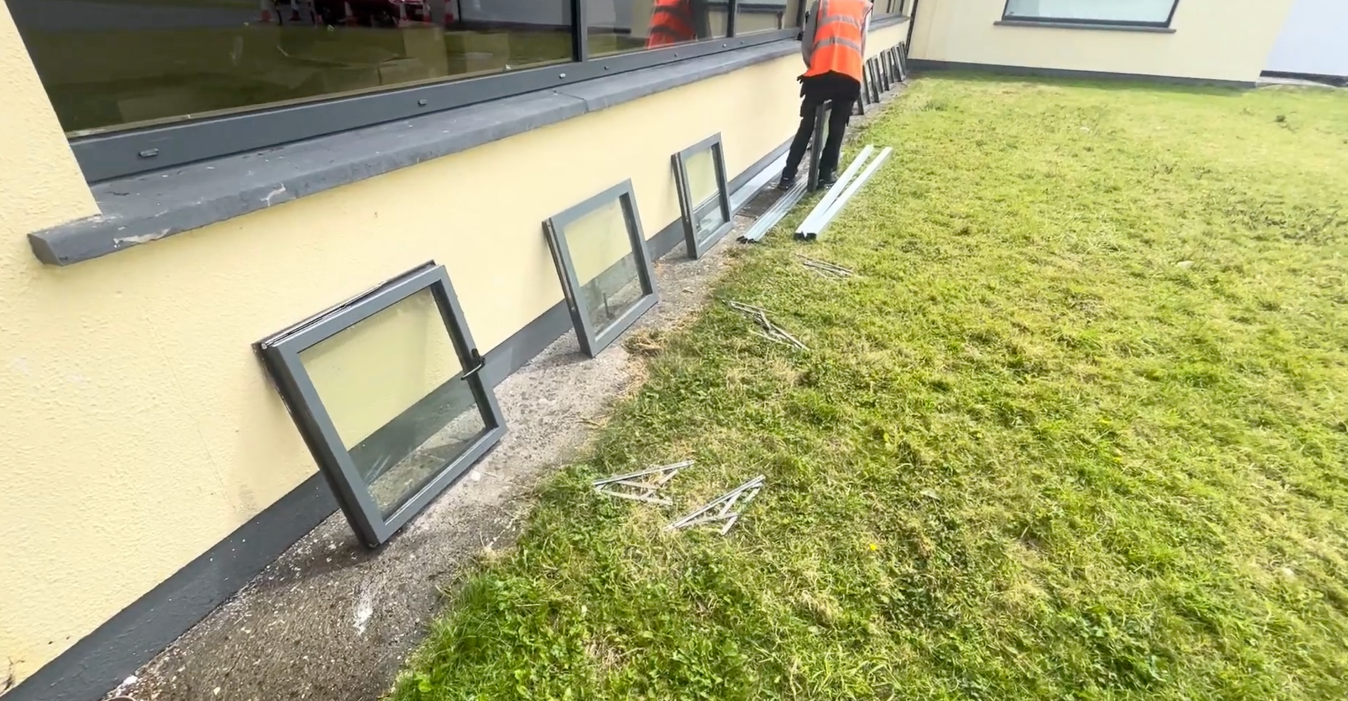 Basement windows set into a sloped grassy yard beside a building, with a worker in orange at the far end.