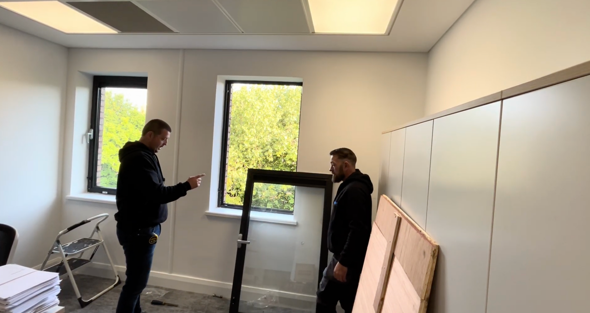 Two people talking in a bright room with large windows and stacked boards against the wall.