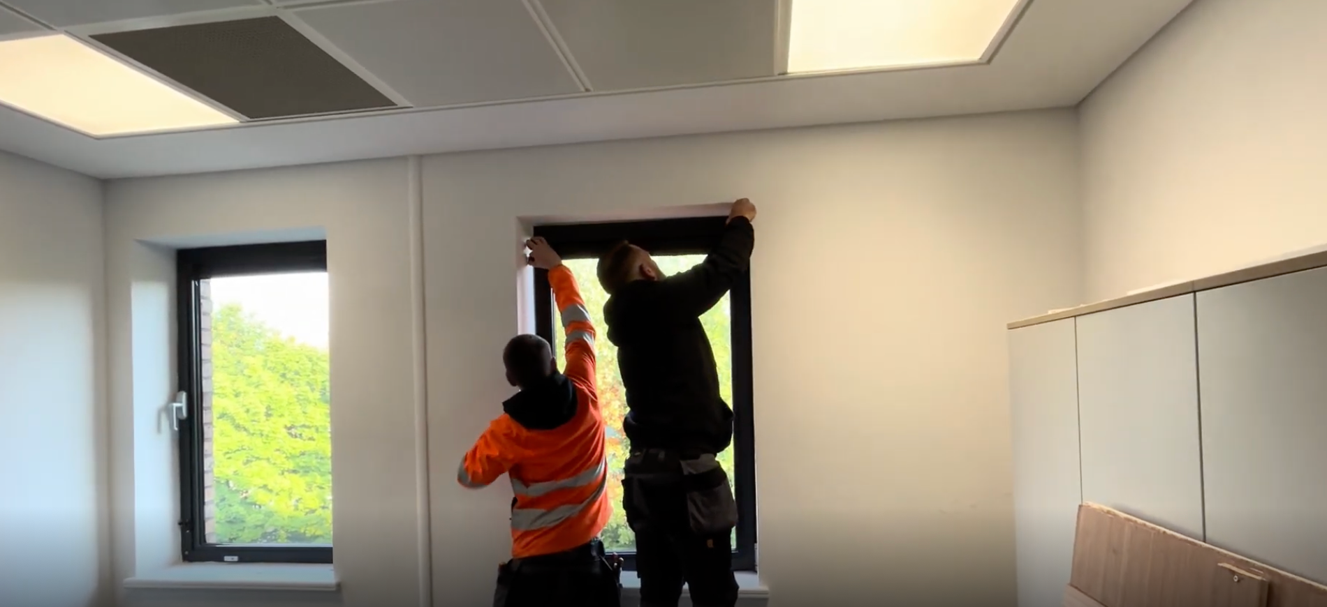 Two workers installing a window blind in a bright room with white cabinets and a window.