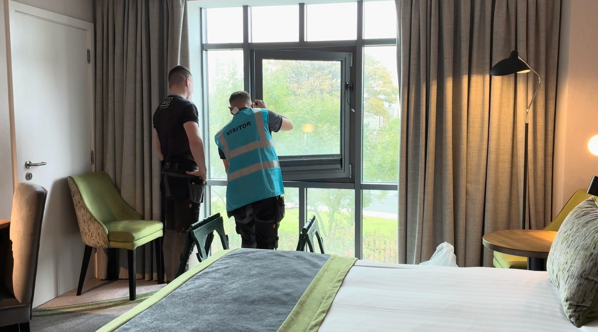 Two people standing by a large window in a hotel room with a bed and curtains.