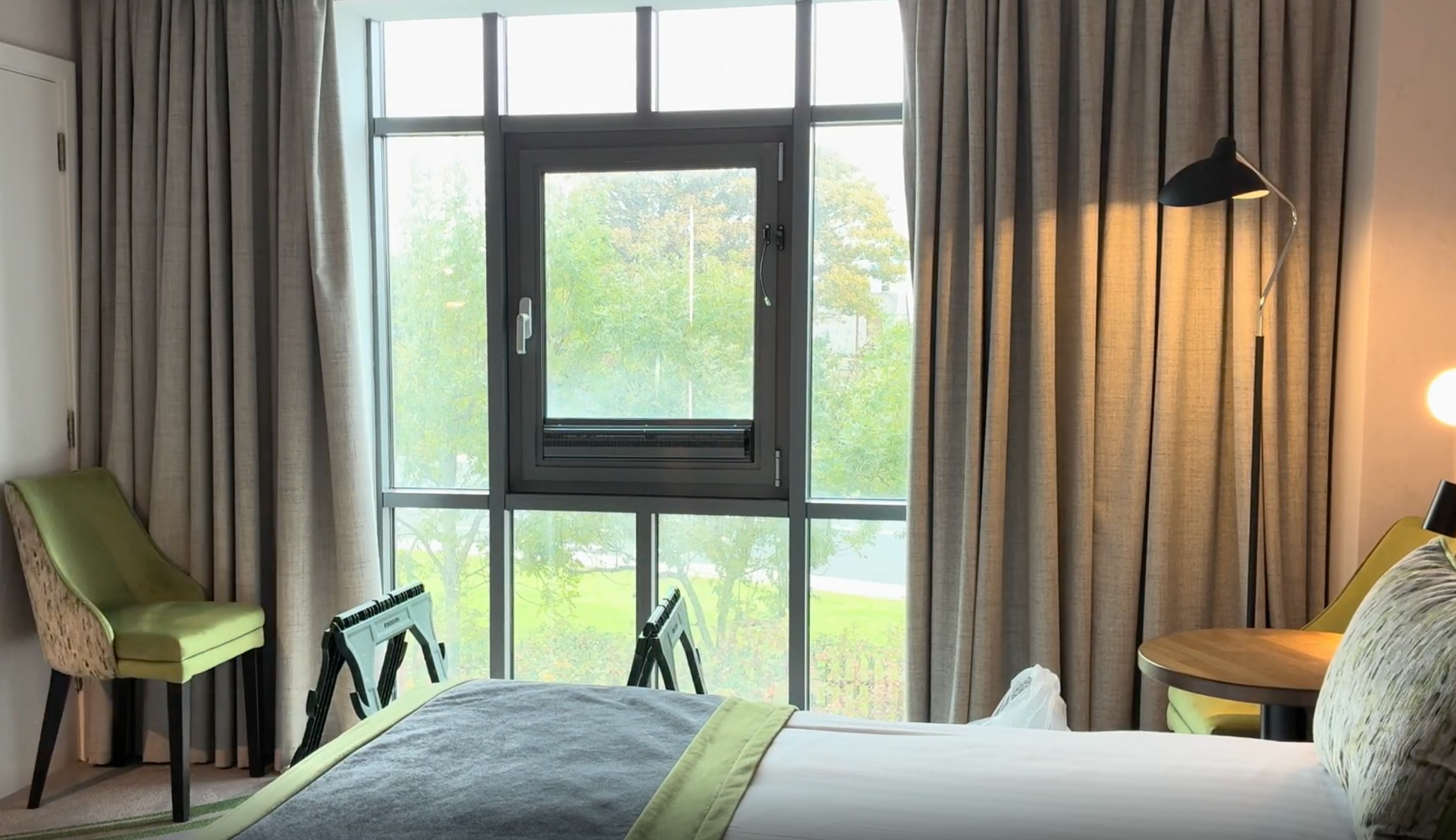 Cozy bedroom with large window, green chair, beige curtains, and warm lamp light