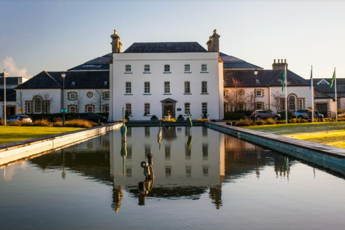 Grand white building reflected in a calm canal at sunset, with flagpoles and landscaped banks