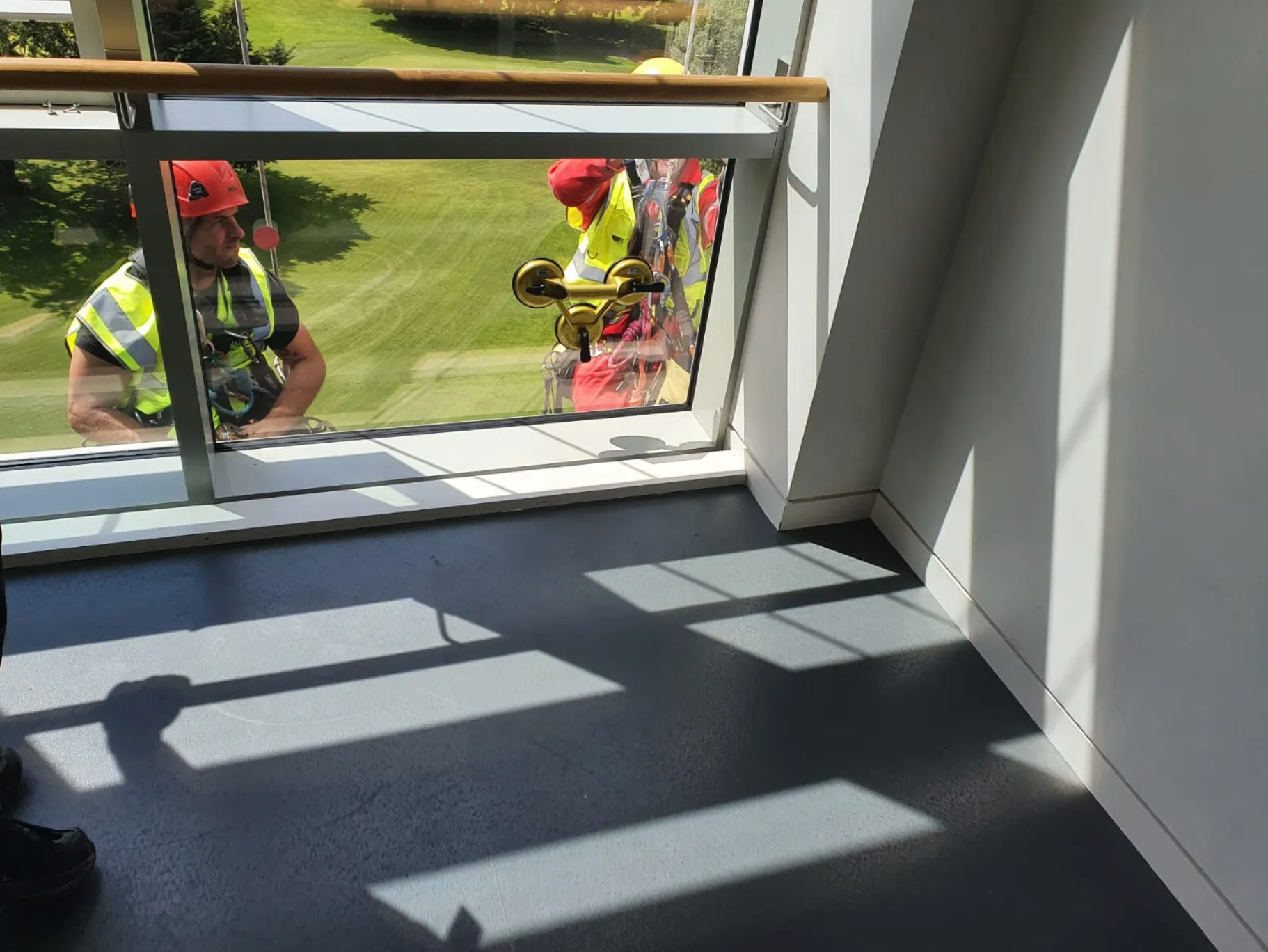 Two workers in yellow vests and red helmets outside a large window, with sunlight casting shadows inside.