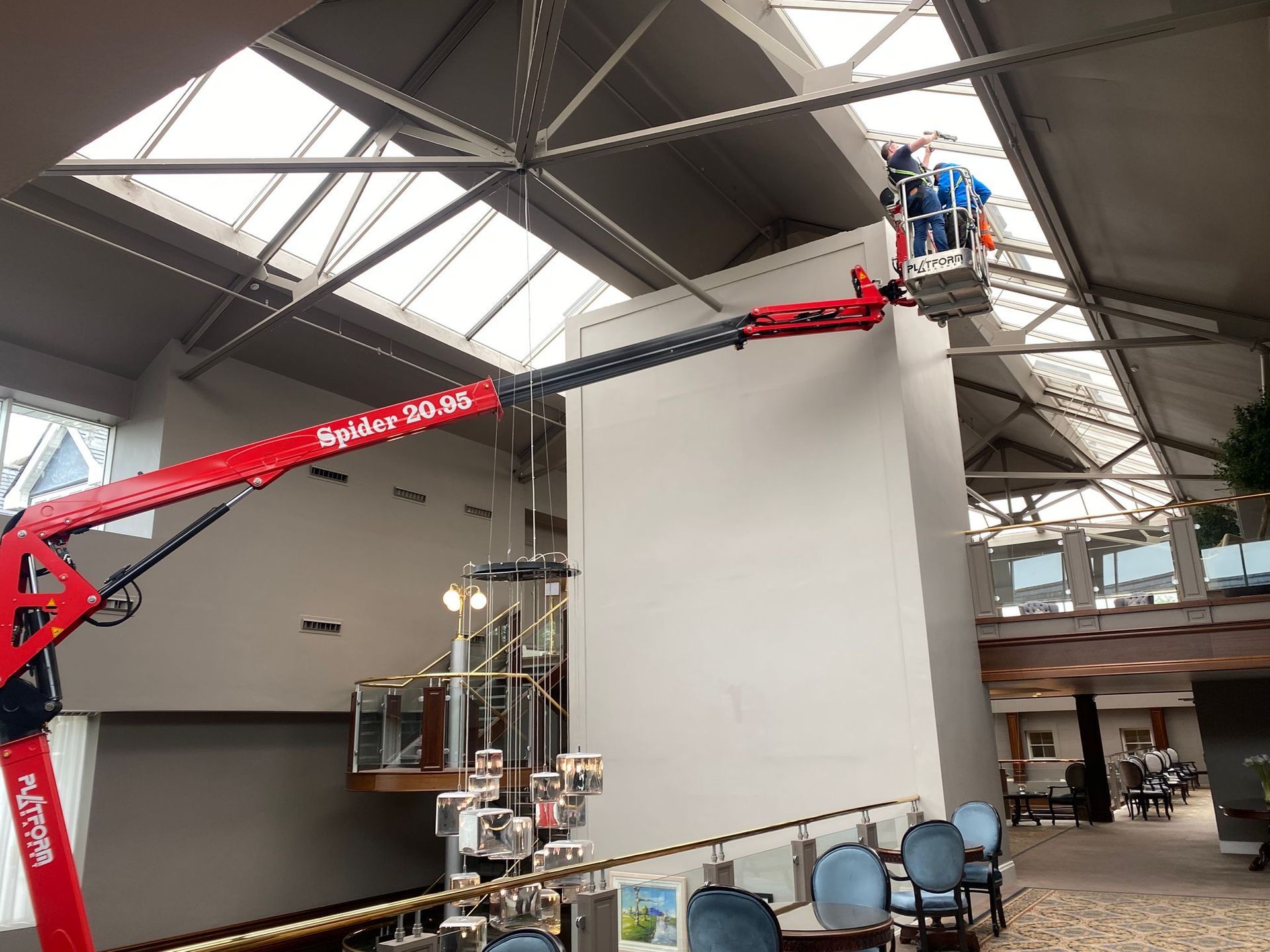 Red boom lift with workers installing equipment on a large white wall in a high-ceilinged atrium.