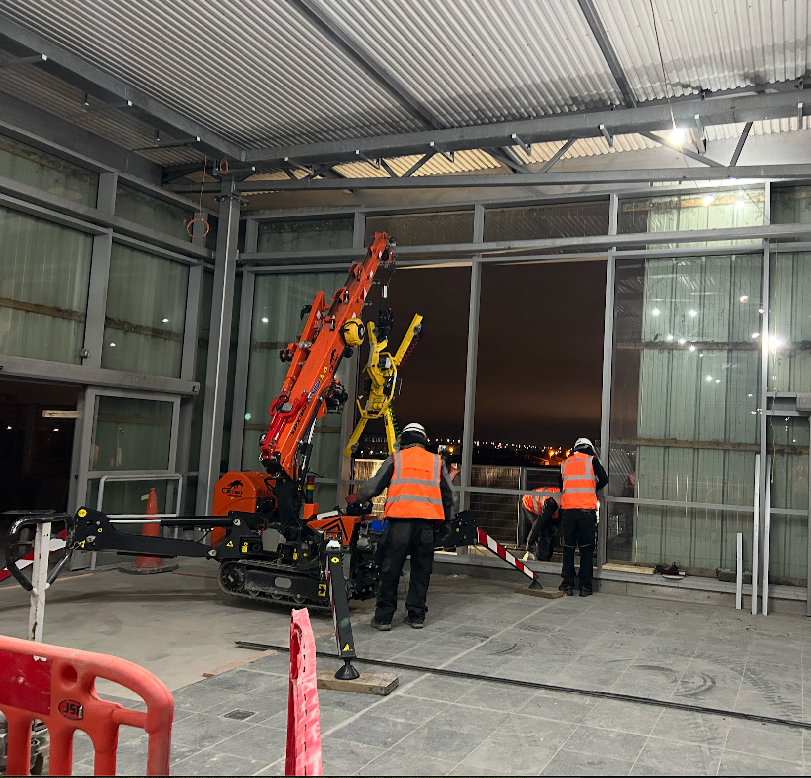 Workers operating orange and yellow lift rigs inside a metal-framed industrial building at night