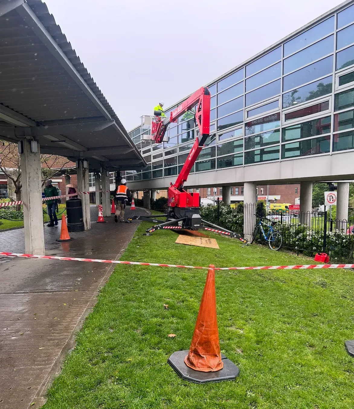 Red cherry picker lifting a worker beside a glass building, with caution tape and orange cones in front.