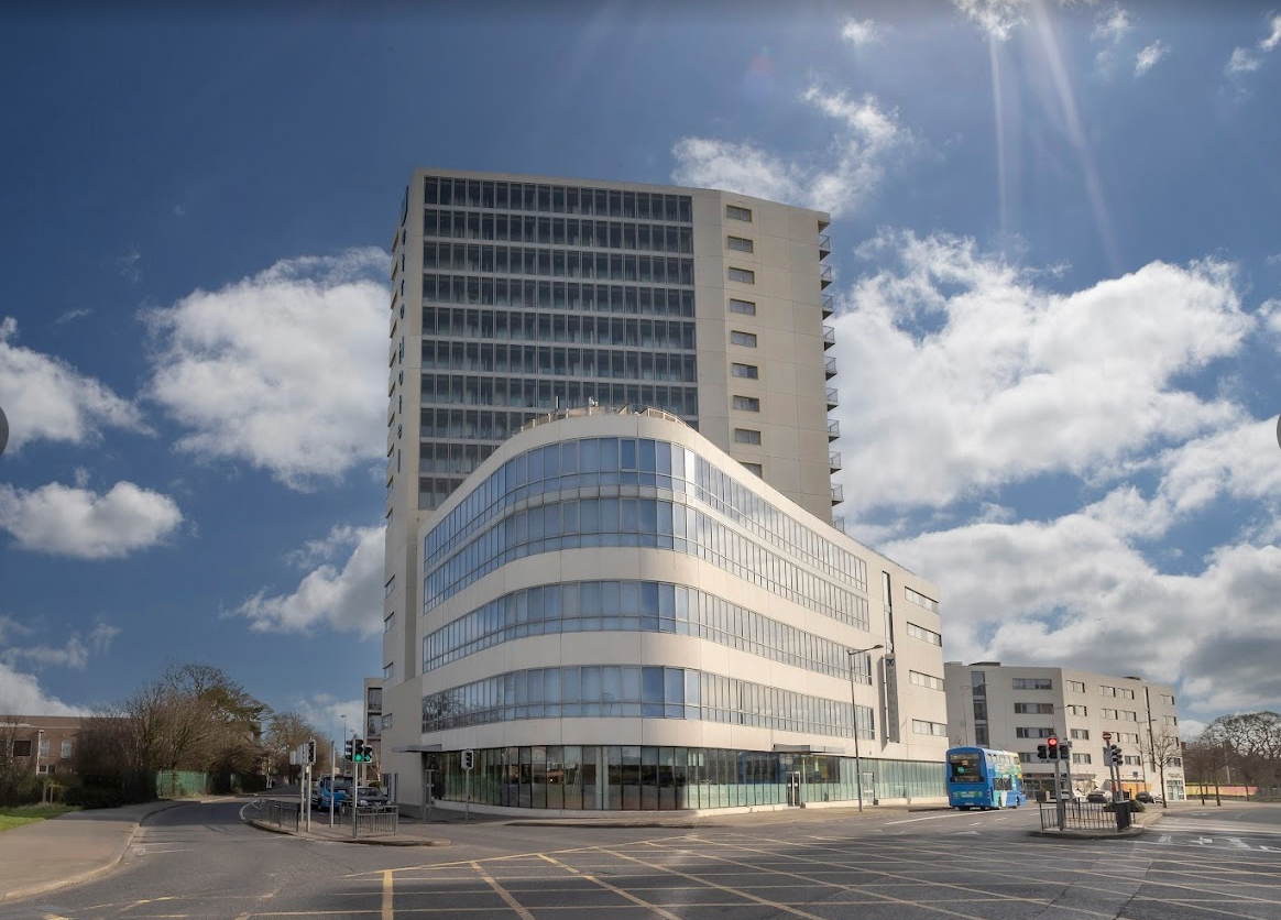 Curved white office building with tall tower behind, seen across an empty street under a blue sky.