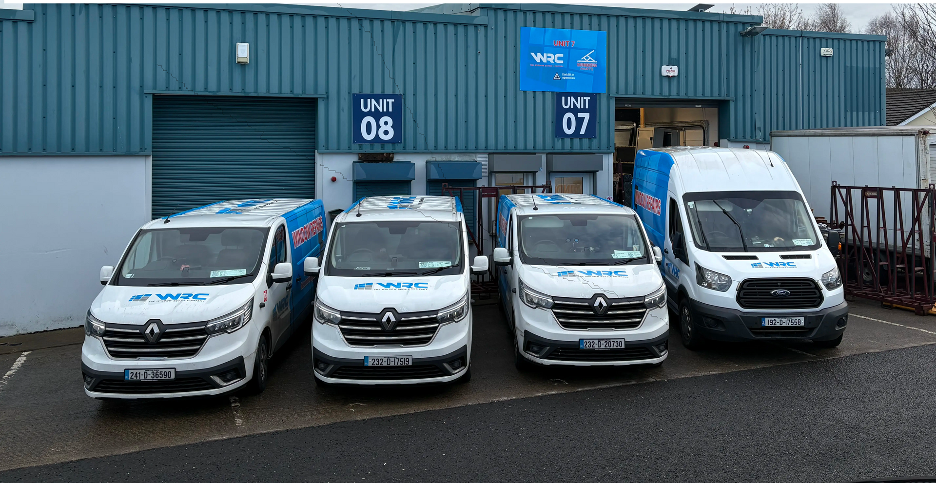 Four parked white service vans in front of a blue industrial building with garage doors.