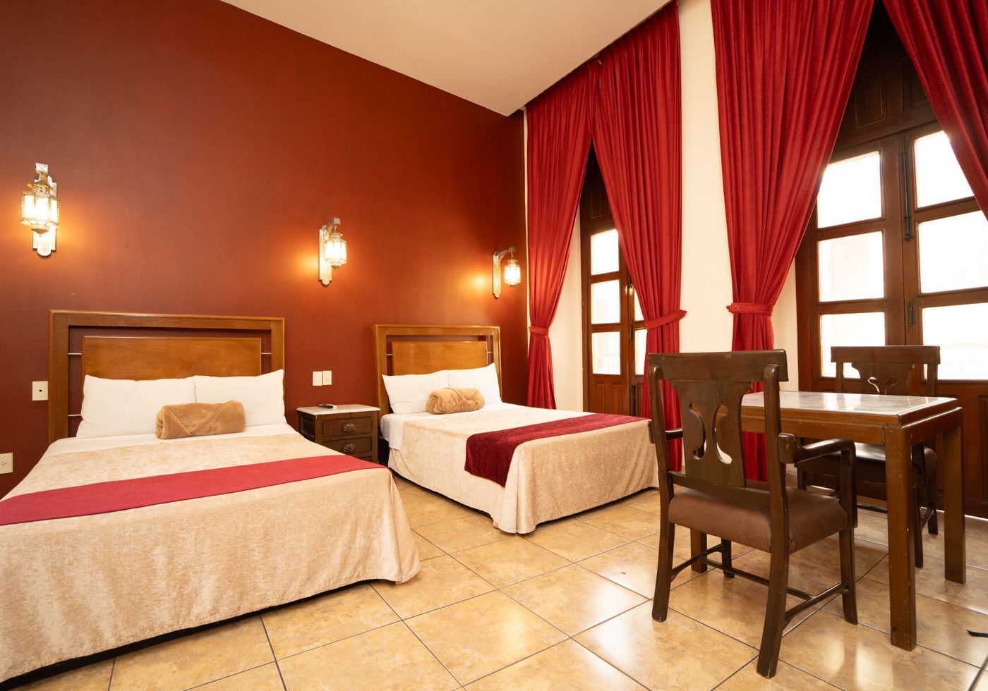 Habitación de hotel con dos camas individuales, paredes y cortinas rojas, suelo de baldosas y una mesita con sillas