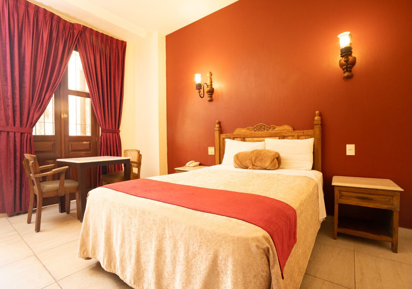 Acogedora habitación de hotel con una cama doble, una pared roja de acento, una mesita de noche y una ventana con cortinas