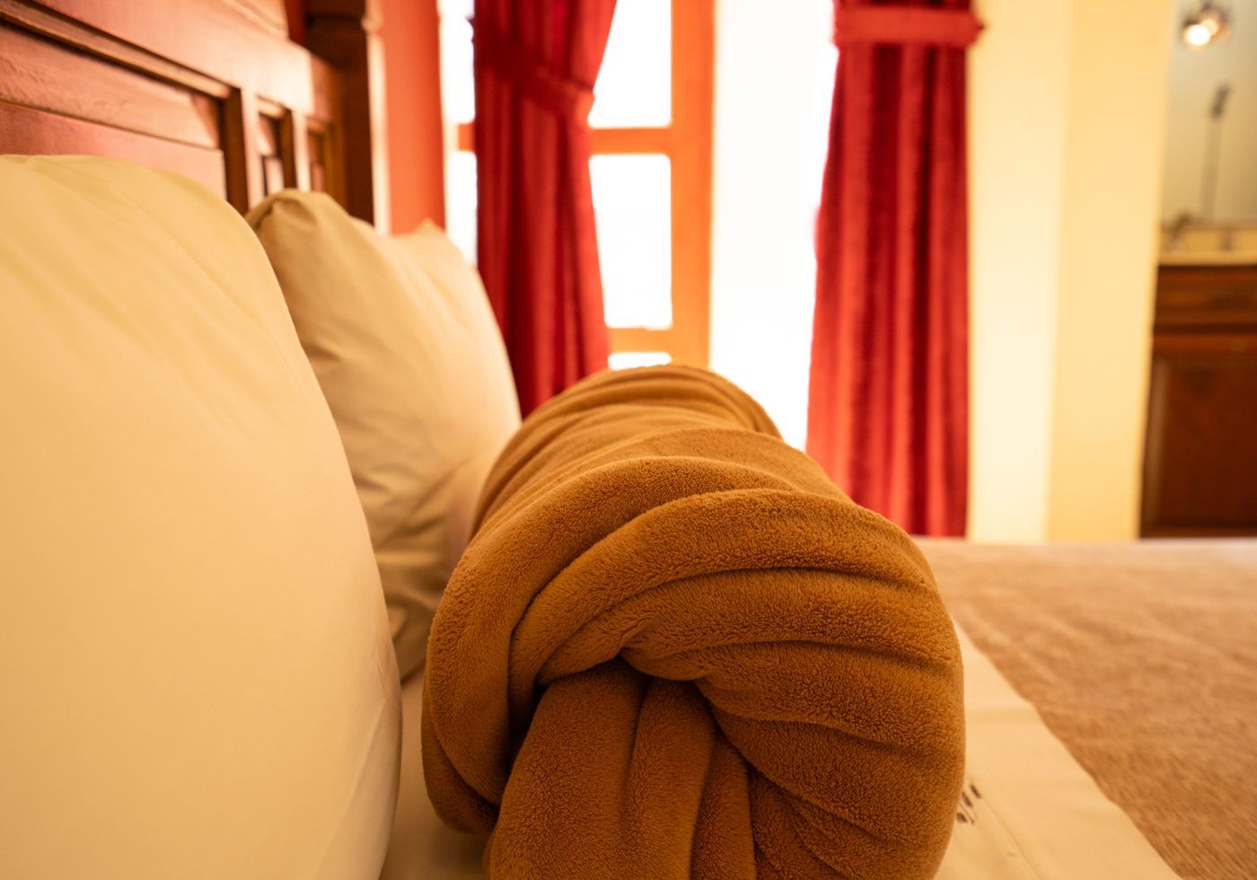 Toalla marrón doblada cuidadosamente sobre una cama en una habitación de hotel soleada con cortinas rojas