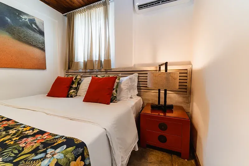 Um quarto com uma cama de casal queen-size com travesseiros vermelhos e um caminho de mesa floral, ao lado de uma mesa de cabeceira vermelha com um abajur.