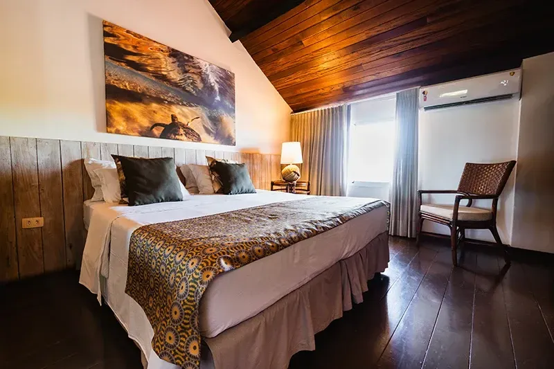 Um quarto de hotel com uma cama grande, tapete estampado, paredes de madeira, teto abobadado e uma cadeira junto à janela.