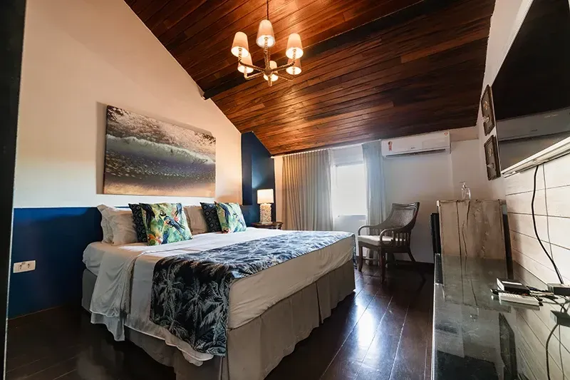 Um quarto de hotel com uma cama grande, teto abobadado de madeira, lustre, parede com detalhes em azul, obras de arte com temática litorânea e uma poltrona.
