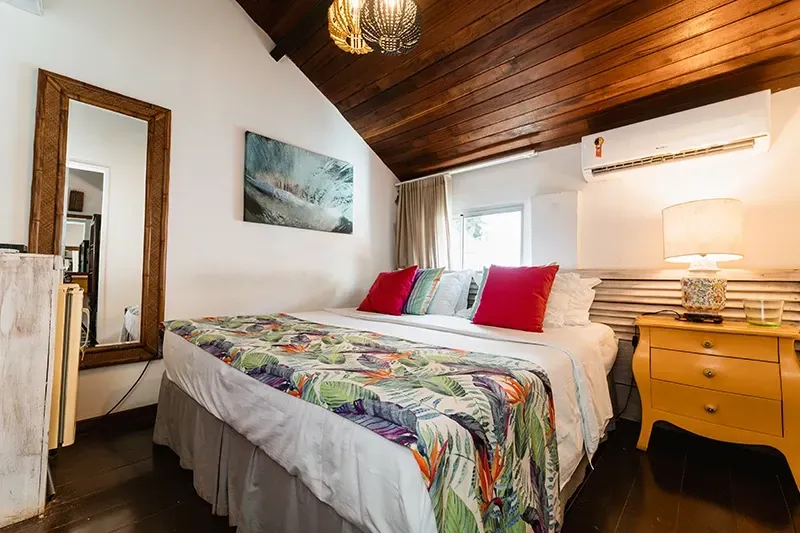 Um quarto com uma cama de casal queen-size, colcha com estampa tropical, travesseiros vermelhos, mesa de cabeceira amarela e teto de madeira.