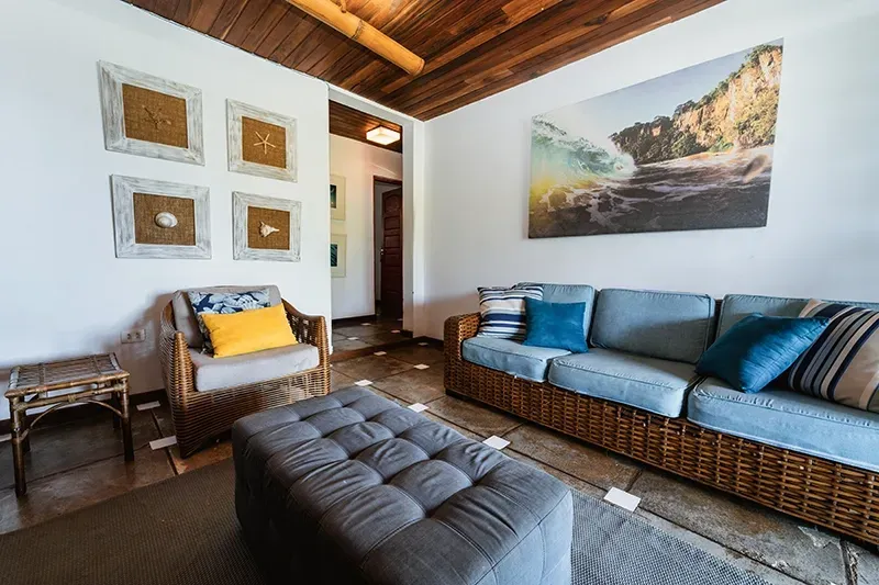 Uma sala de estar com sofá e poltrona de vime, pufe cinza capitonê, obras de arte com tema de praia na parede e teto com ripas de madeira.