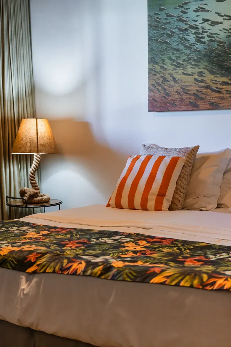 Um quarto de hotel com uma cama com uma manta de estampa tropical e uma almofada listrada laranja ao lado de um abajur em uma mesa de cabeceira.