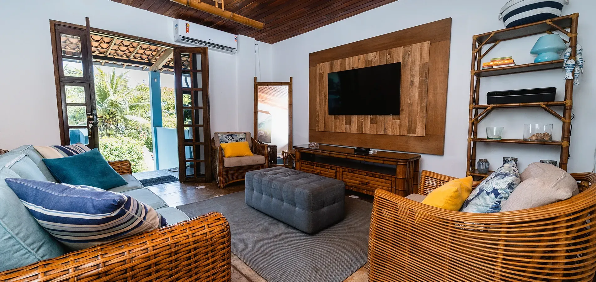 Uma sala de estar aconchegante com móveis de vime, um pufe cinza, uma TV fixada na parede com painéis de madeira e portas de vidro que dão para um pátio.