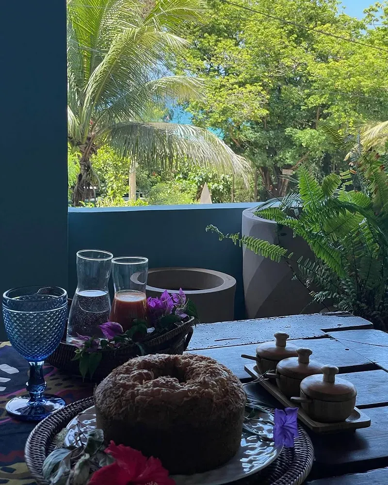 Um bolo redondo repousa sobre uma mesa de madeira ao lado de copos, tigelas pequenas e flores roxas, com exuberantes árvores tropicais ao fundo.