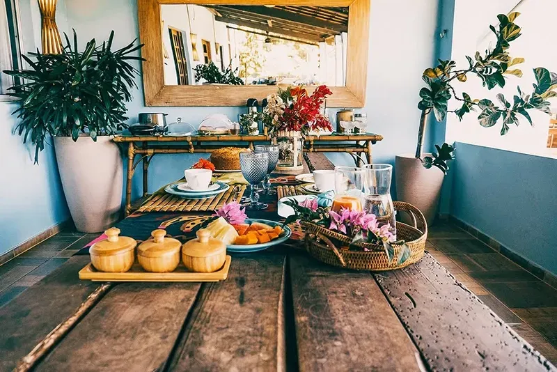 Uma mesa rústica de madeira posta para uma refeição em uma varanda ensolarada, com pequenos recipientes, flores e plantas exuberantes em vasos.