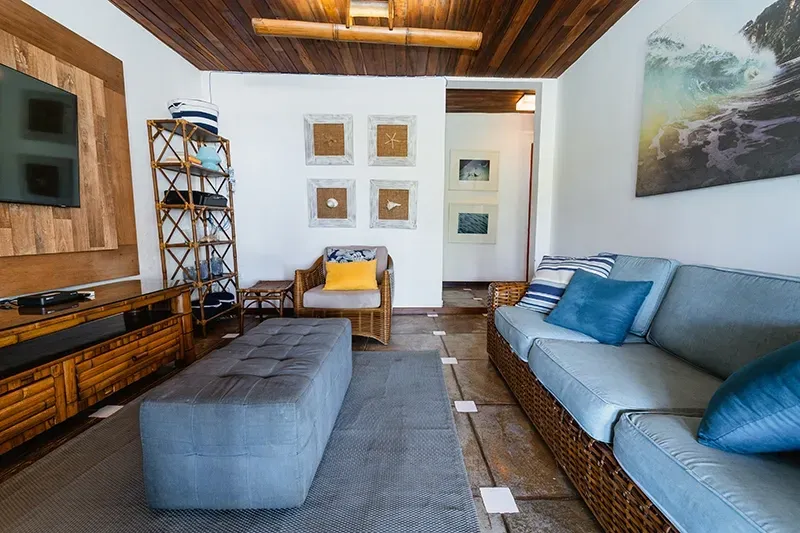 Sala de estar com sofá azul, pufe combinando, poltrona de vime, estante de madeira, TV e teto estilo bambu.