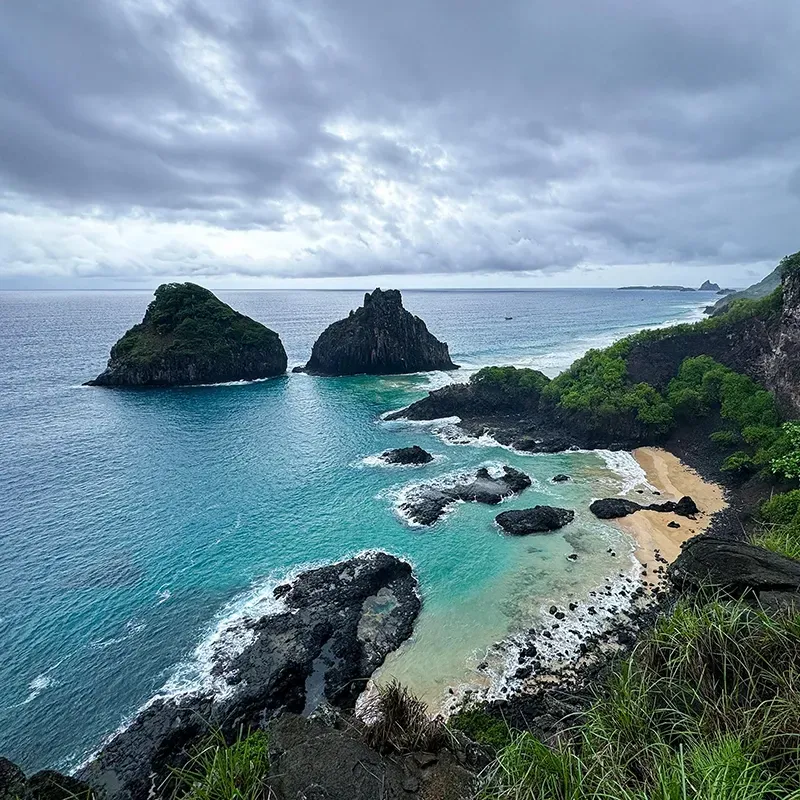 Vista aérea da costa rochosa de Fernando de Noronha, com águas turquesa, enseadas de areia e falésias verdejantes.