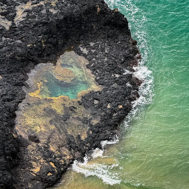 Uma poça de maré turquesa se encontra em uma rocha vulcânica negra e acidentada, ao lado de um oceano turquesa com ondas brancas quebrando.