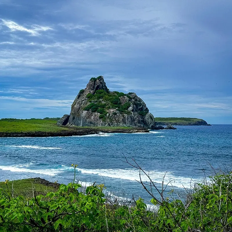 Uma grande formação rochosa escarpada emerge do oceano azul, rodeada por colinas costeiras verdejantes sob um céu nublado.