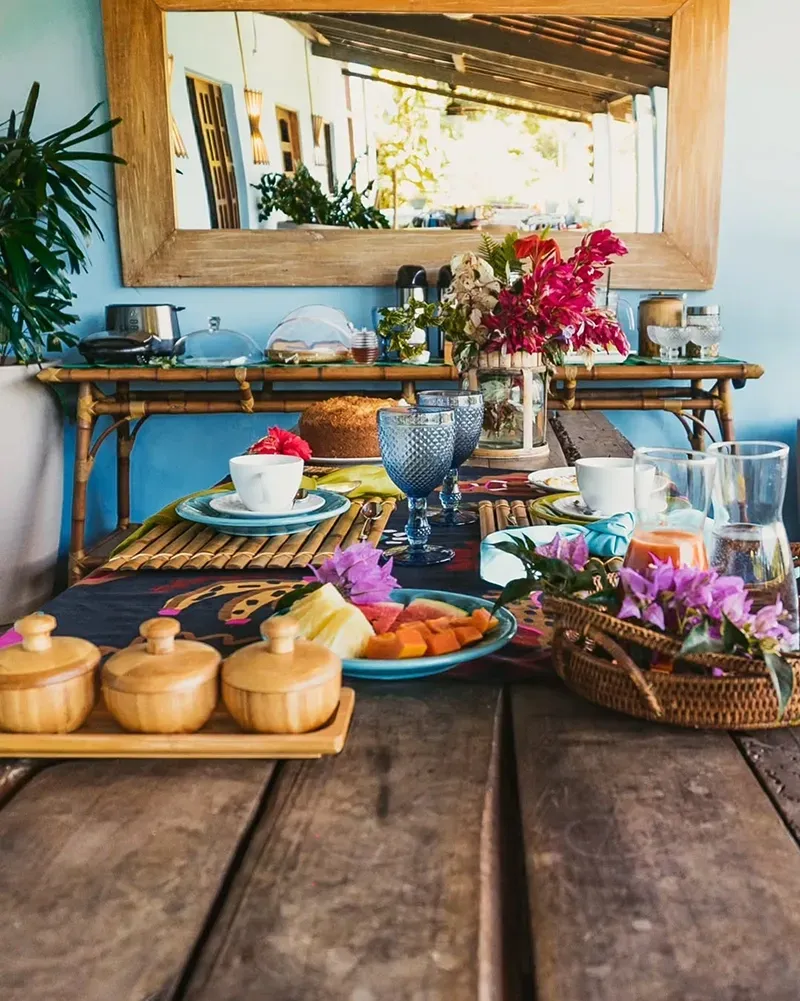Uma mesa rústica de madeira posta para o café da manhã com um bolo, frutas frescas, xícaras, um arranjo de flores e um console espelhado.