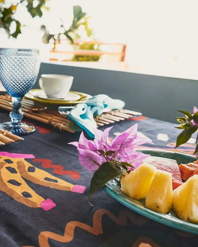 Uma toalha de mesa estampada com um copo azul, uma xícara de café e um prato com abacaxi e melão, decorado com uma flor roxa.