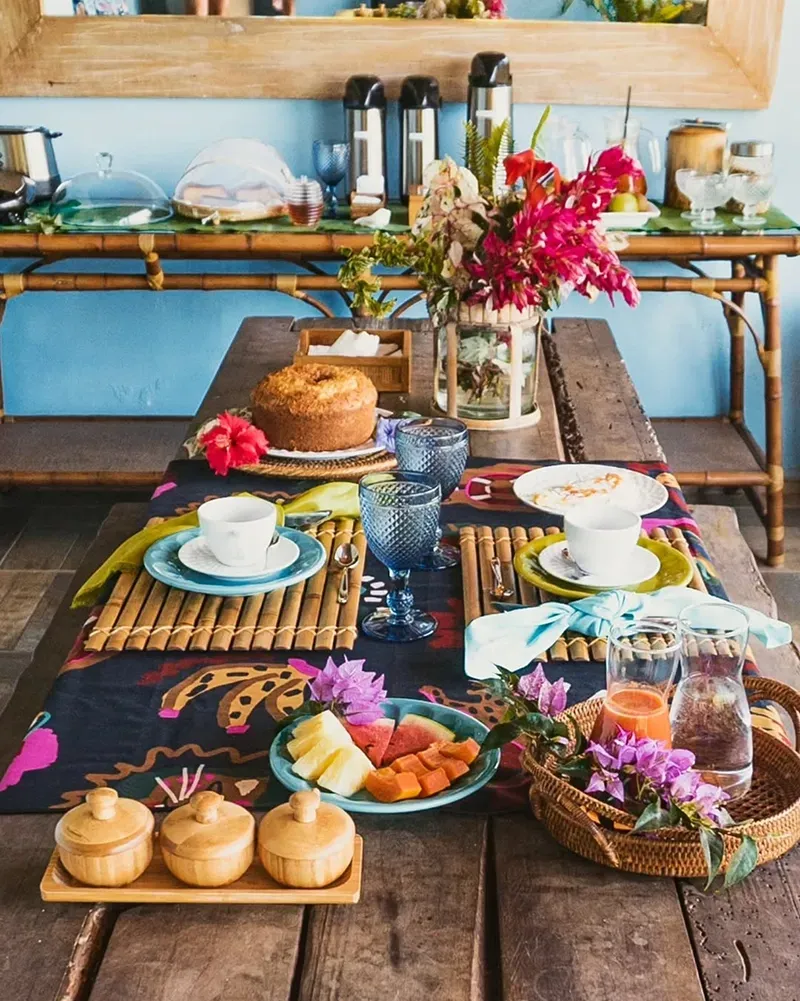 Uma mesa posta para um café da manhã tropical com frutas, bolo, xícaras de chá e arranjos florais em uma sala de jantar rústica.